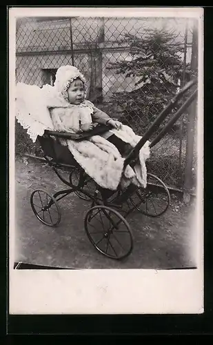 Foto-AK Kind mit Mütze im Kinderwagen vor einem Gartenzaun