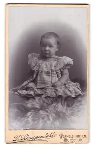 Fotografie G. Schneppendahl, Wermelskirchen, Kleines Kind im karierten Kleid sitzt auf einem Fell