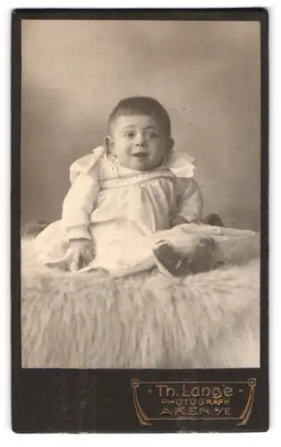 Fotografie Th. Lange, Aken a. E., Süsses Kleinkind im Kleid sitzt auf einem Fell