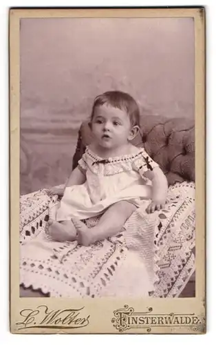 Fotografie L. Wolters, Finsterwalde, Grabenstrasse 4, niedliches Baby im weissen Kleidchen auf Spitzendecke sitzend