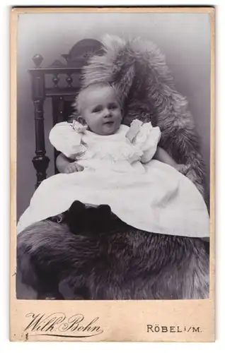 Fotografie Wilh. Bohn, Röbel i. M., Kirchplatz & Stavenstr., niedliches kleines Kind im weissen Kleid auf Fell sitzend