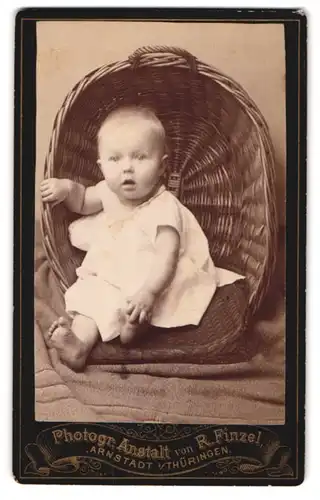 Fotografie R. Finzel, Arnstadt i. Th., Kleinkind im weissen Leibchen sitzt in einem Weidenkorb