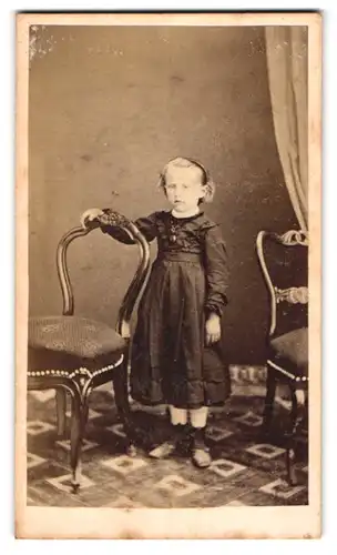 Fotografie unbekannter Fotograf und Ort, niedliches blondes Mädchen im dunklen Kleid steht zwischen den Stühlen