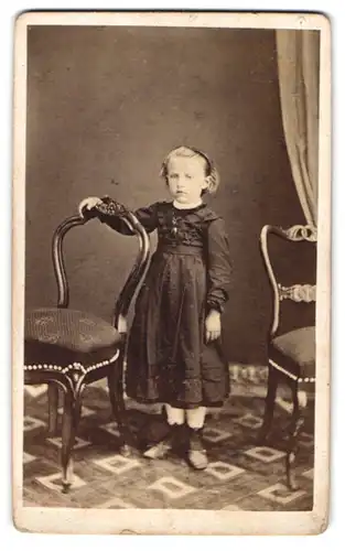 Fotografie unbekannter Fotograf und Ort, kleines blondes Mädchen im schwarzen Kleid steht zwischen den Stühlen