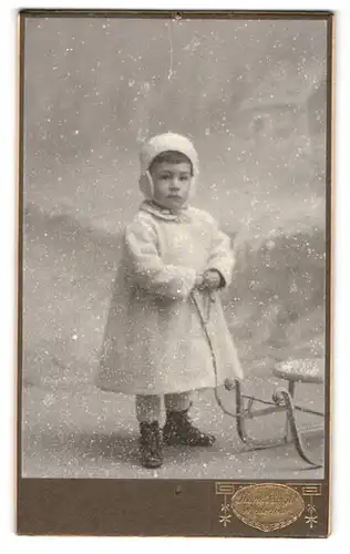 Fotografie Hans Jäggli, Winterthur, niedliches Kind im Winterkleid mit Wintermütze zieht den Schlitten, Studiokulisse