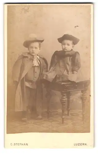 Fotografie C. Stephan, Luzern, zwei Kleinkinder im Anzug mit Mantel und im Kleidchen auf Tisch sitzend