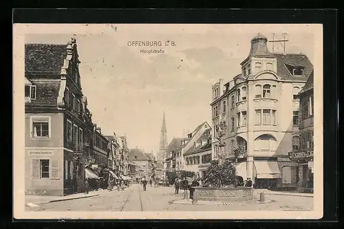 AK Offenburg i. B., Hauptstrasse mit Geschäften, Kirchturm und Burnnen