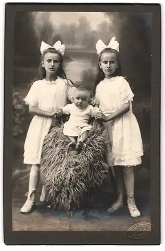 Fotografie Eugen Hofbauer, Suhl, zwei niedliche Schwestern in weissen Kleidern mit ihrem Geschwisterkind auf Fell sitzend