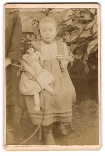 Fotografie unbekannter Fotograf und Ort, kleines Mädchen im Kleid mit grosser Puppe im Arm