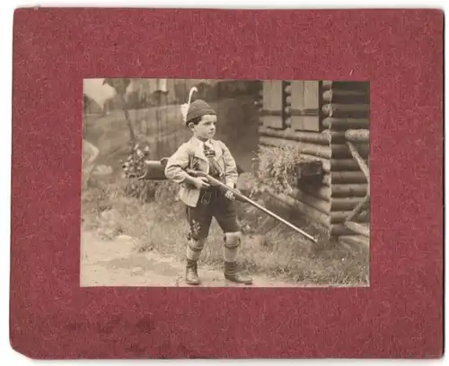 Fotografie unbekannter Fotograf und Ort, junger Knabe in Lederhosen mit Scheibenbüchse vor einer Studiokulisse