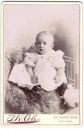 Fotografie J. K. Cole, New York, Sixth Ave. 174, süsser kleiner Knabe Conrad Neumann im weissen Kleidchen auf Fell sitzend