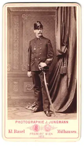 Fotografie J. Jungmann, Kleinbasel, Portrait schweizer Artillerist in Uniform mit Säbel und Tschako, Ärmelabzeichen