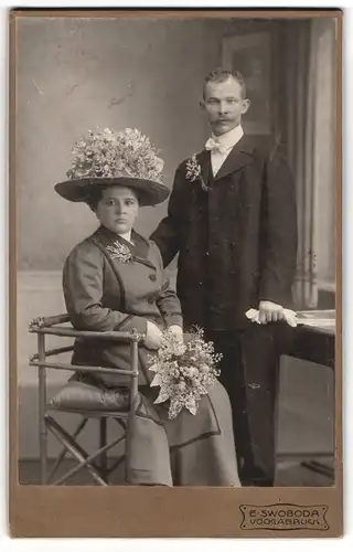 Fotografie E. Swoboda, Vöcklabruck, Ehepaar im dunklen Kleid mit Blumenhut und Mann im Anzug