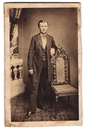 Fotografie unbekannter Fotograf und Ort, junger Mann im Anzug mit Fliege stehend neben einem Stuhl