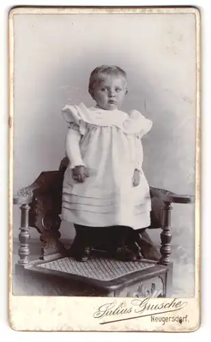 Fotografie Julius Grusche, Neugersdorf, niedliches Kleinkind im weissen Kleidchen stehend auf einem Stuhl