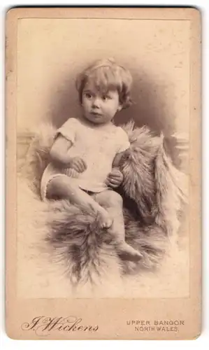 Fotografie J. Wickens, Bangor, niedliches Kleinkind im Wollkleid sitzt auf Fell