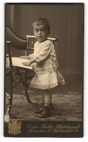 Fotografie Oswin Fiedler, Dresden, junger Knabe im Kleidchen stehend am Stuhl