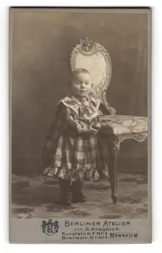 Fotografie E. Kregeloh, Mannheim, Kunststr. Nr. 3, Süsses Kleinkind mit abstehenden Ohren am Stuhl stehend