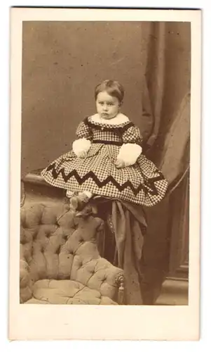 Fotografie unbekannter Fotograf und Ort, niedliches kleines Mädchen im karierten Kleid sitzt auf der Sessellehne