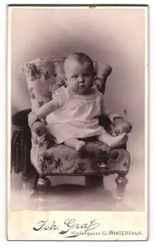 Fotografie Joh. Graf, Winterthur, Niedergasse 13, Niedliches Kleinkind auf Stuhl sitzend im weissen Kleidchen