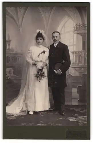 Fotografie Fritz Hacker, Annaberg, Carlsplatz 3, Eheleute im Brautkleid und Anzug mit Zylinder vor einer Studiokulisse