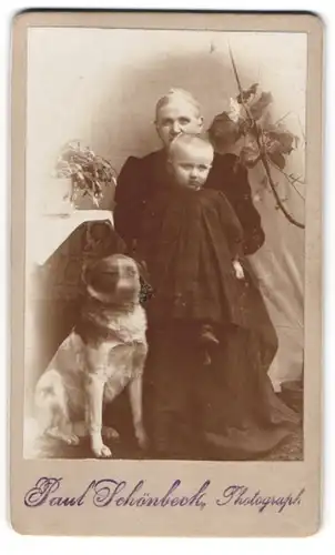 Fotografie Paul Schönbeck, Ort unbekannt, Grossmutter mit Enkelkind auf dem Schoss und Hund an der Seite