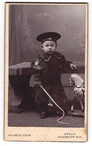 Fotografie Wilhelm Stein, Berlin, Kleinkind im dunklen Kleid mit Spielzeug Pferd und Peitsche