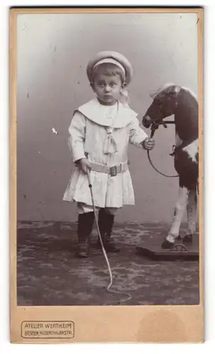 Fotografie Wertheim, Berlin, junger Knabe im weissen Kleid mit Schauklepferd und Peitsche