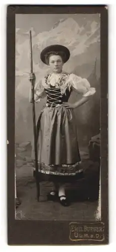 Fotografie Emil Burger, Ulm a. D., junge Frau im Trachtenkleid mit Wanderstock vor einer Studiokulisse
