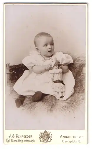 Fotografie J. S. Schroeder, Annaberg i. S., niedliches Kleinkind mit seiner Puppe auf Fell sitzend
