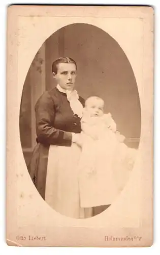 Fotografie Otto Liebert, Holzminden a. W., Junge Frau im Schürzenkleid mit Baby im Taufkleid