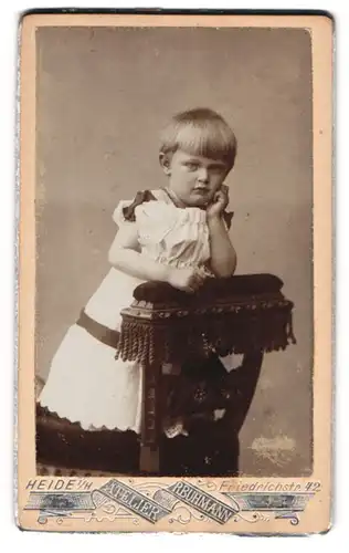 Fotografie Atelier R. Buhmann, Heide /H., Friedrichstrasse 42, Kind im Kleidchen auf einem Stuhl