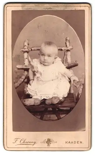 Fotografie F. Chromy, Kaaden, Brunnersdorfergasse 509, Niedliches kleines Baby in weissem Kleid auf grossem Stuhl