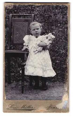 Fotografie Louis Müller, Hameln, niedliches kleines Mädchen mit ihrer Puppe im Arm steht neben einem Stuhl