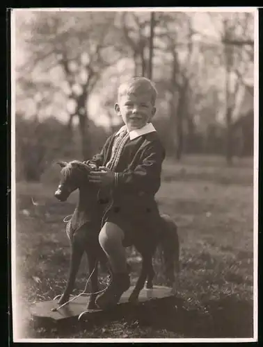 Fotografie blonder lächelnder Knabe auf Spielzeug-Pferd sitzend