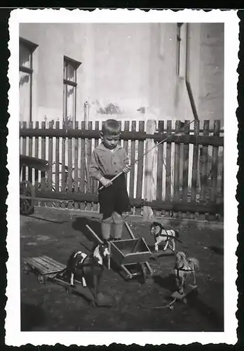 Fotografie Knabe mit Reitgerte & Spielzeug-Pferden im Garten