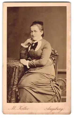 Fotografie M. Keller, Augsburg, Zeuggasse 226b, Elegante Dame mit Spitzenkragen und Medaillon, an einem Tisch sitzend