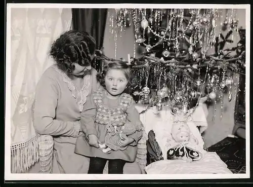 Fotografie Weihnachten, staunendes Mädchen nebst Puppe vor Weihnachtsbaum sitzend