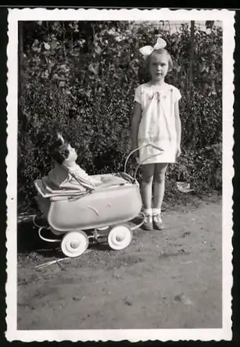 Fotografie Mädchen mit Haarschleife nebst Puppe im Puppenwagen
