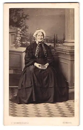 Fotografie J. Schneebeli, Zürich-Niederdorf, betagte Dame mit Haube vor Studiokulisse sitzend