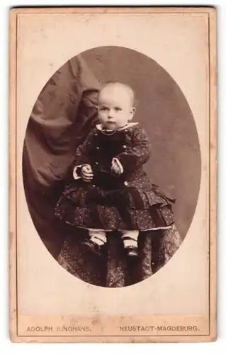 Fotografie Adolph Junghans, Neustadt-Magdeburg, Kleinkind in dunklem Kleid auf Tisch sitzend