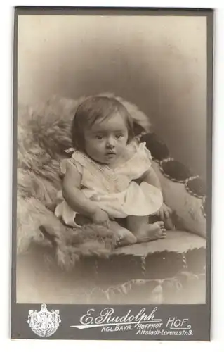 Fotografie E. Rudolph, Hof, Portrait sitzendes Kleinkind im weissen Hemd mit nackigen Füssen