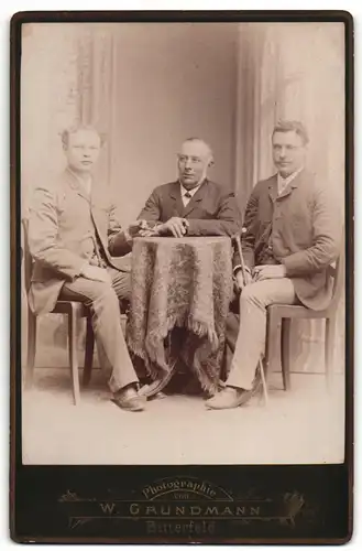 Fotografie W. Grundmann, Bitterfeld, Portrait drei modisch gekleidete Herren mit Zigarren am Tisch