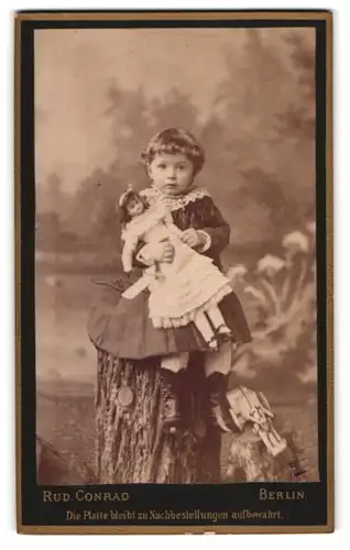 Fotografie Rud. Conrad, Berlin, süsses kleines Mädchen mit ihrer Puppe, sitzend auf Baumstamm
