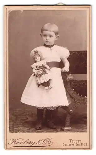 Fotografie Kanberg & Co., Tilsit, Mädchen im weissen Kleid mit Puppe im Arm, Topfhaarschnitt