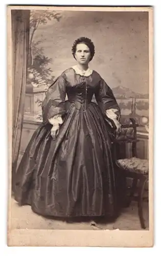 Fotografie P. C. Schiller, Frankfurt / Main, Dame im dunklen Reifrock Kleid mit Haube vor einer Studiokulisse