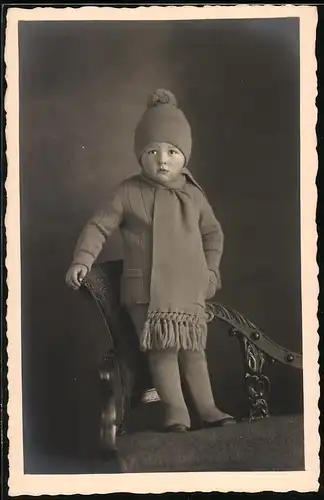 Fotografie Billström, Kiel, Kleinkind in Winterbekleidung auf Sessel stehend