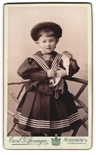 Fotografie Carl G. Springer, Reichenberg i. B., niedliches kleines Mädchen im Matrosenkleid mit Puppe im Arm