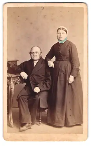 Fotografie B. Sparmeyer, Herrnhut, älteres Paar im dunklen Kleid mit Haube und im Anzug sitzend am Sekretär