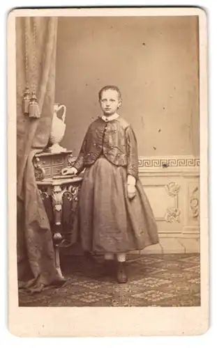 Fotografie F. A. Wilde, Weissenfels, junges blondes Mädchen im schlichten Kleid stehend am Sekretär